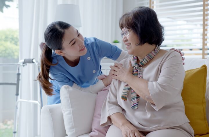 image of a young asian nurse putting her hand on the shoulder of an older asian woman who is sitting on a chair inside a home