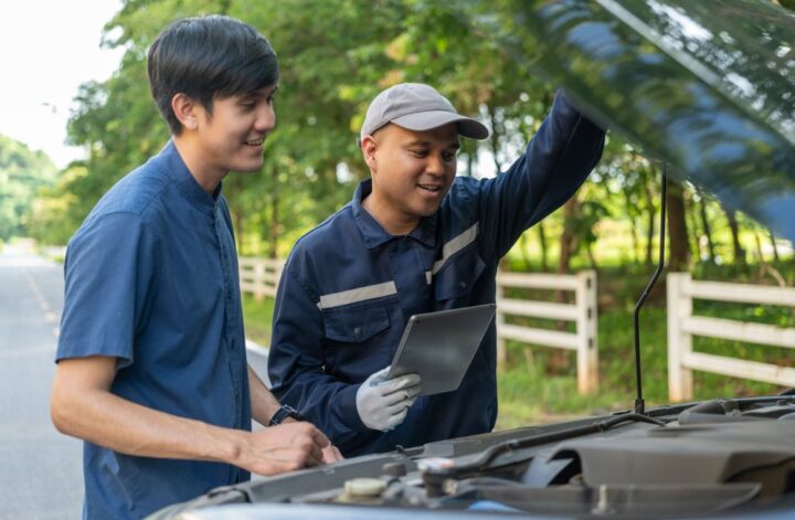 Two men stand by a car with its hood open—one holding a tablet and wearing gloves—outdoors on a roadside with trees and a fence in the background, as they review what a transmission specialist checks during repairs.